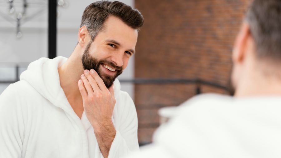 man applying beard conditioner on his beard while facing the mirror